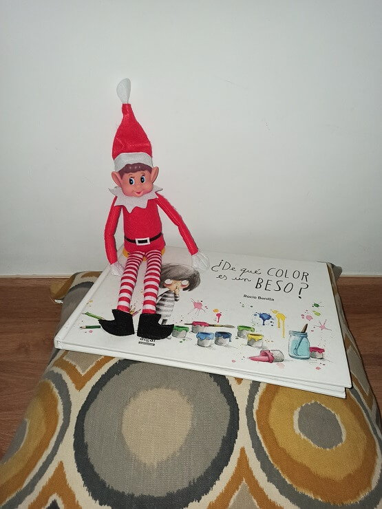 El elfo de Navidad con el libro ¿De qué color es un beso? de Rocío Bonilla. El elfo en el estante